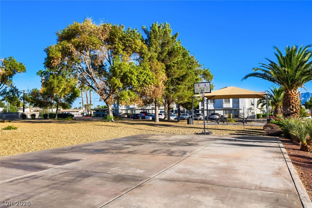 A spacious outdoor recreation area features a concrete basketball court surrounded by mature shade trees.