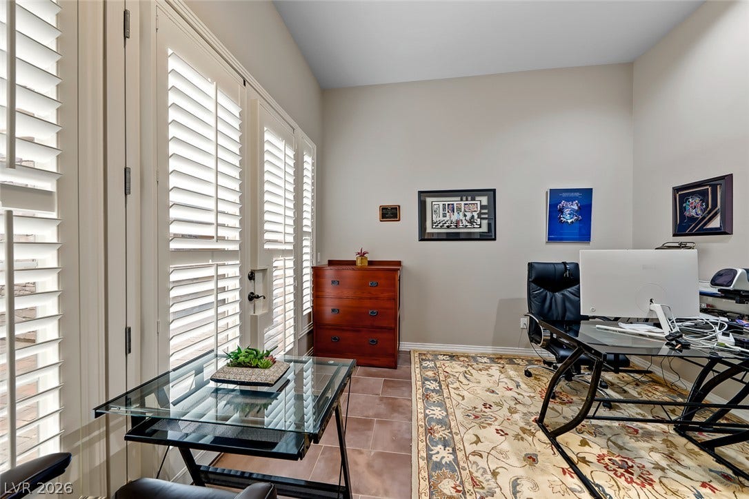 Sunlight streams through plantation shutters into a quiet home office with tiled flooring.