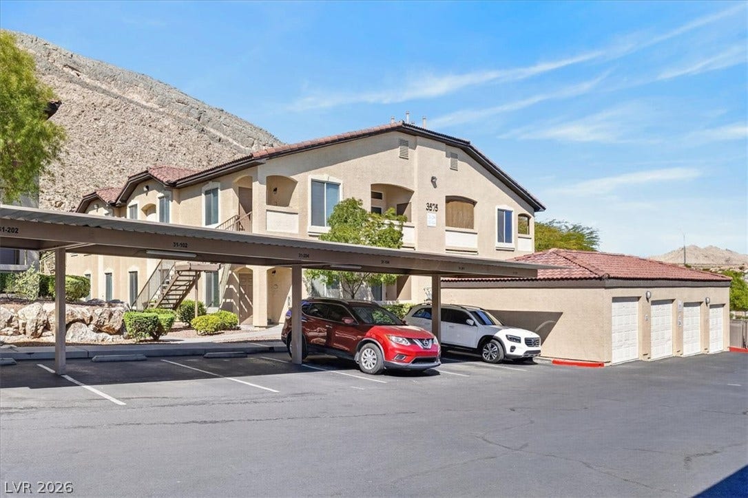 A multi-level stucco building features covered parking spaces against a desert mountain backdrop.