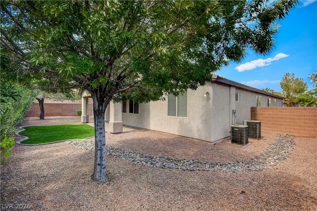 Mature trees shade a tidy gravel yard next to a stucco home with a covered patio.