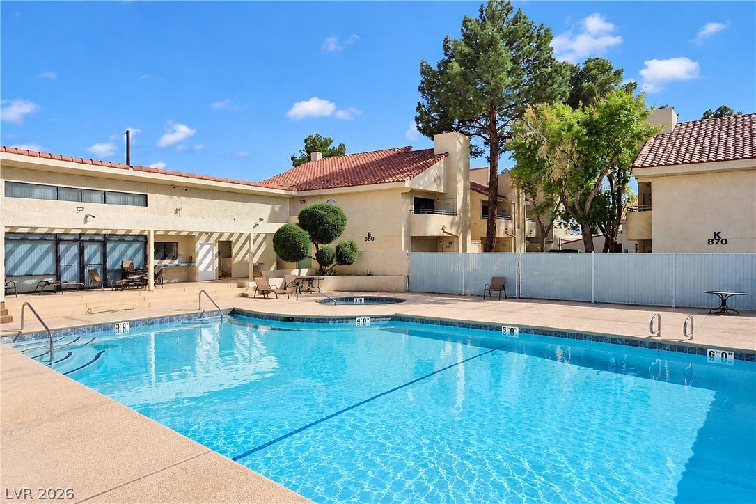 A sparkling outdoor pool is surrounded by a spacious concrete patio and stucco buildings.