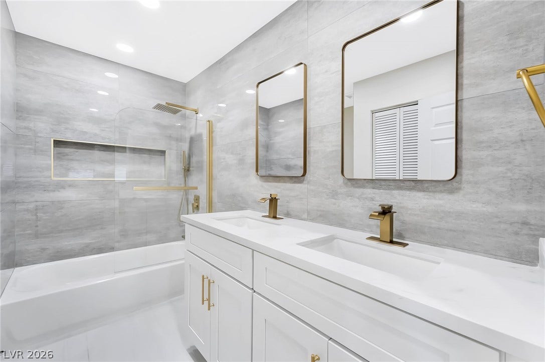 Gold fixtures accent this dual-sink vanity alongside a glass-enclosed tub with floor-to-ceiling tiling.
