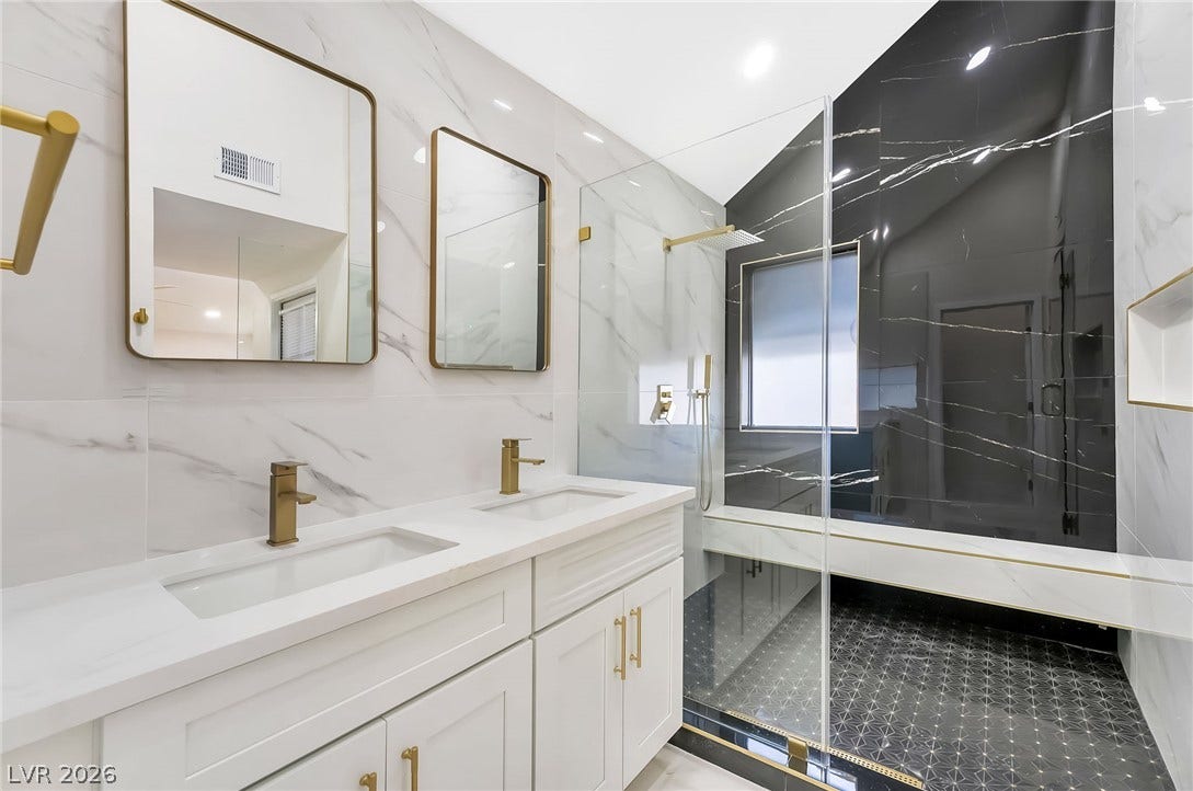Gold fixtures accent a sleek double vanity and a glass-enclosed shower with custom tile.