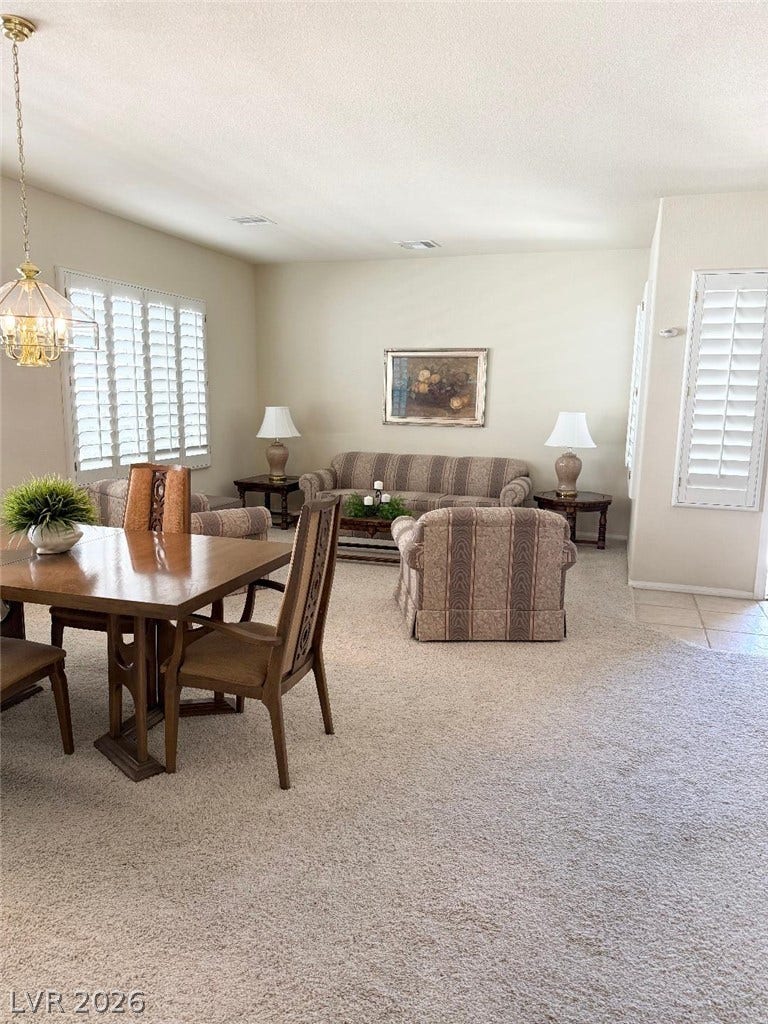 An open living and dining area features neutral carpeting, plantation shutters, and tall ceilings.