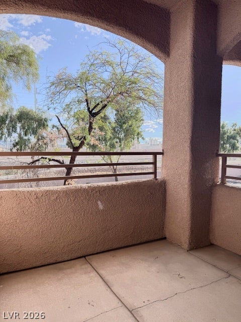 An arched covered balcony looks out toward mature trees and a sunny blue sky.