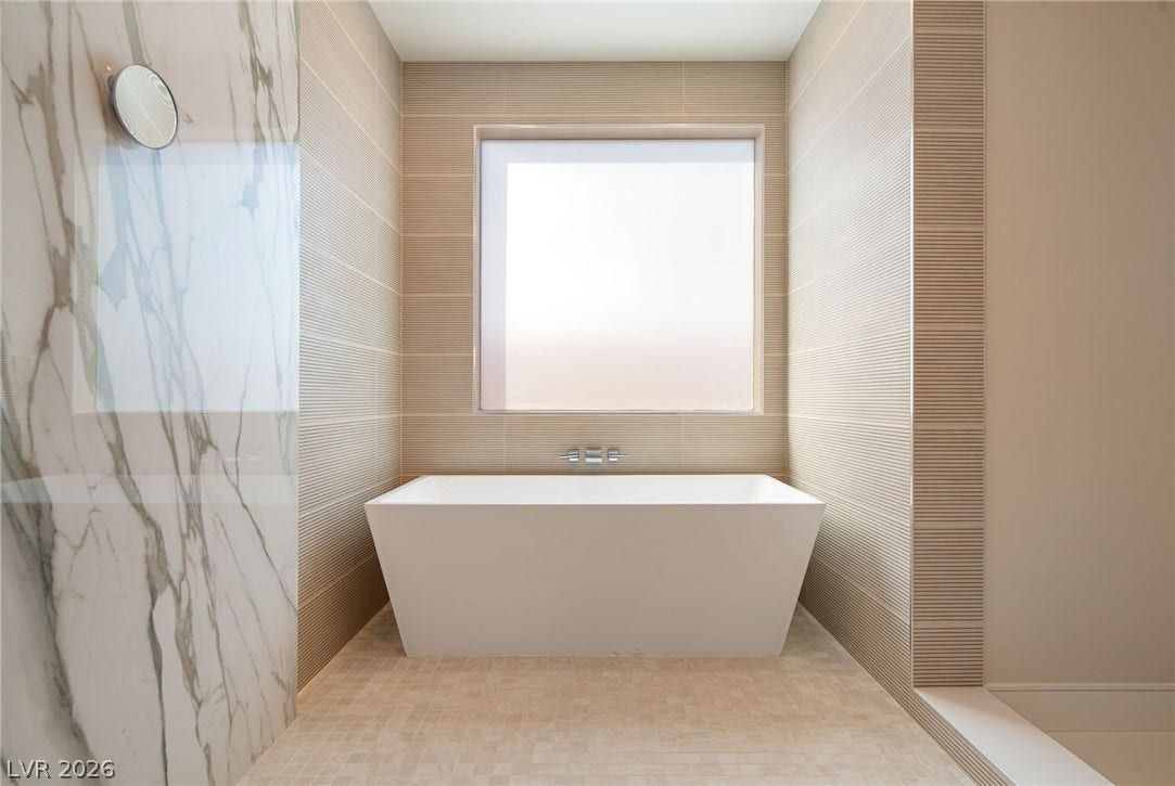 A modern soaking tub rests beneath a large window surrounded by elegant textured tile.
