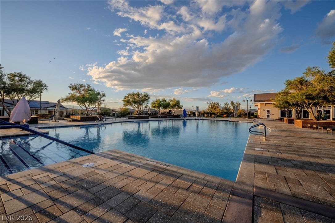 A spacious outdoor community pool is surrounded by extensive stone paver decking and mature trees.