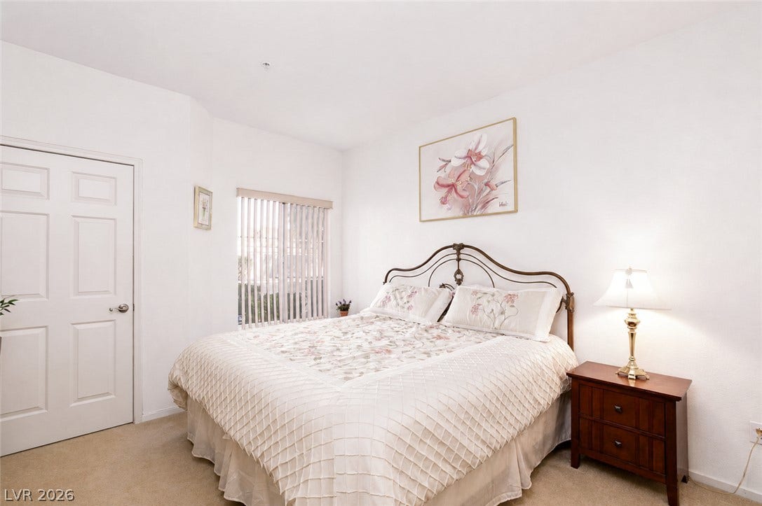 A window with vertical blinds filters natural light across this carpeted bedroom.