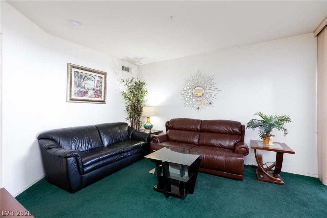 A spacious living area featuring wall-to-wall carpeting and clean white walls.