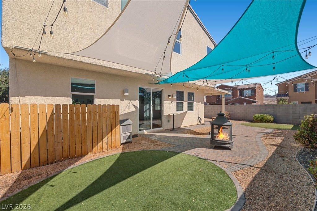 An inviting backyard features paved patio spaces and a warm fire pit under shade sails.