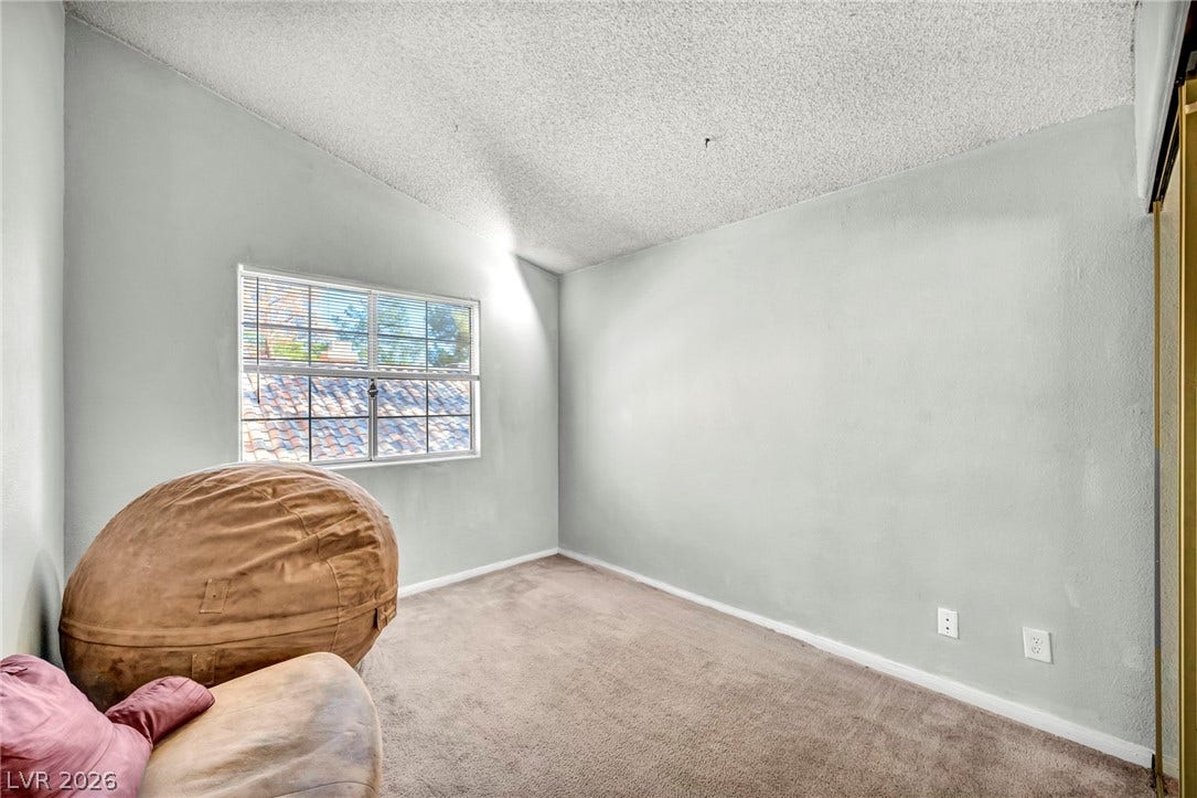 A quiet bedroom features soft carpet flooring, high vaulted ceilings, and a large window.