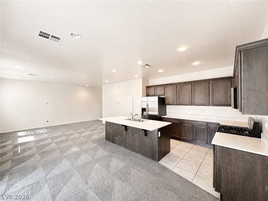 An open kitchen with dark cabinets and a large island overlooks a carpeted living space.