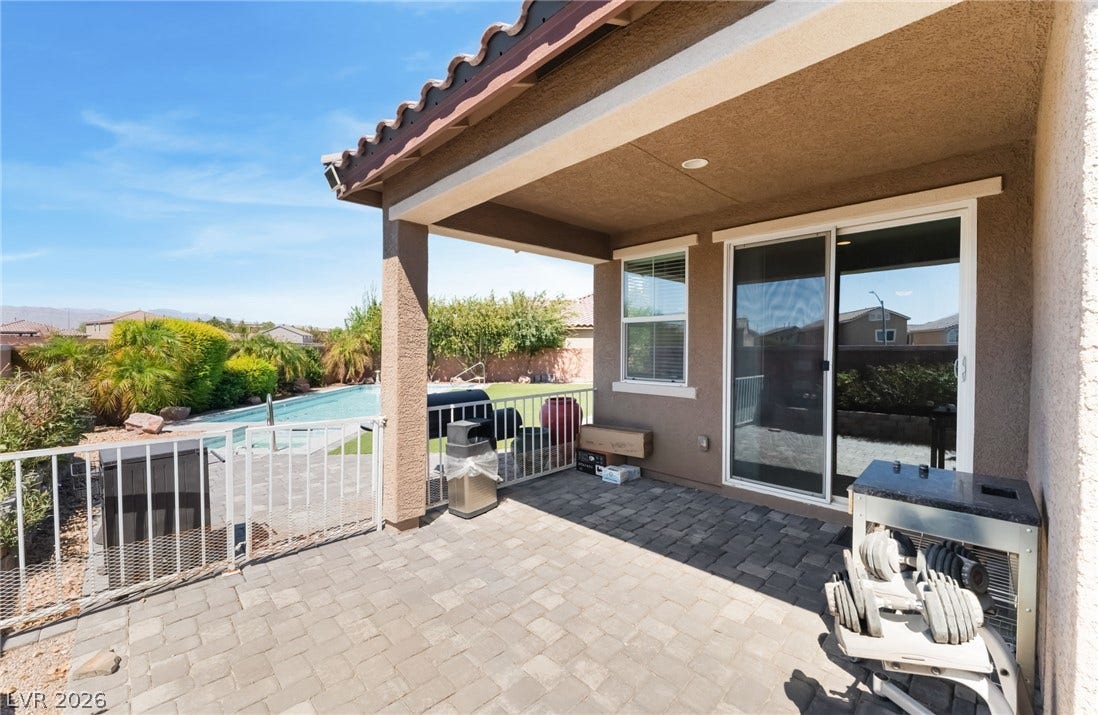Stone pavers line a covered patio overlooking a fenced swimming pool under sunny skies.