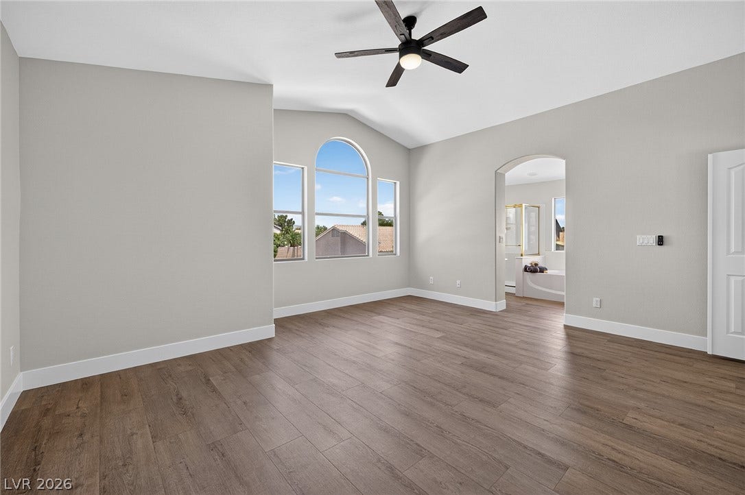 Arched window detail overlooking backyard, ceiling fan and vaulted ceilings define this inviting primary bedroom, seamlessly flowing into a private en-suite bathroom.