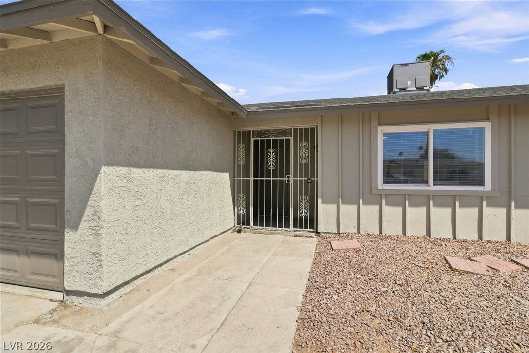 A concrete walkway leads to a secure gated entrance alongside textured siding and gravel landscaping.