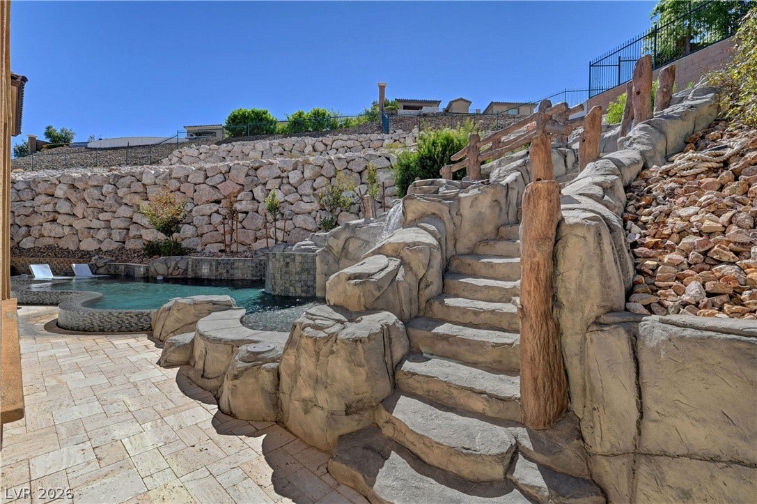 89 Olympia Chase Drive, Las Vegas, NV 89141 Stone steps climb past a waterfall feature toward the tiered, stone-walled backyard landscape.