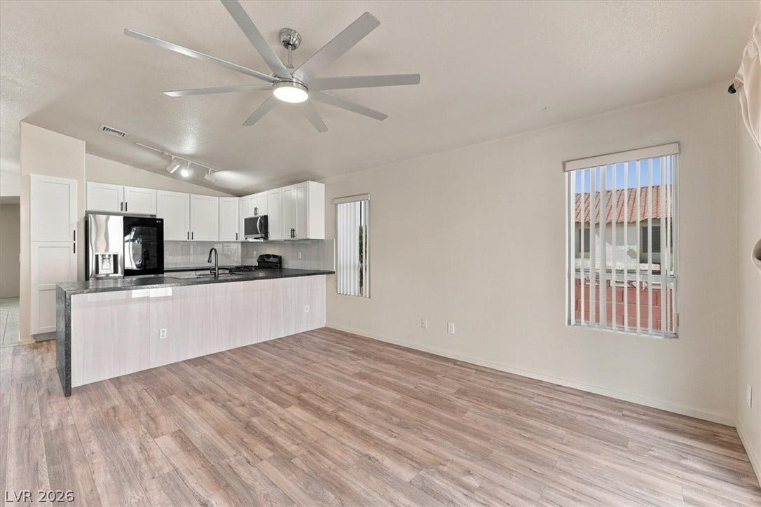 An open living space transitions seamlessly into a modern kitchen with wood-look flooring throughout. Ceiling fan and window covers.