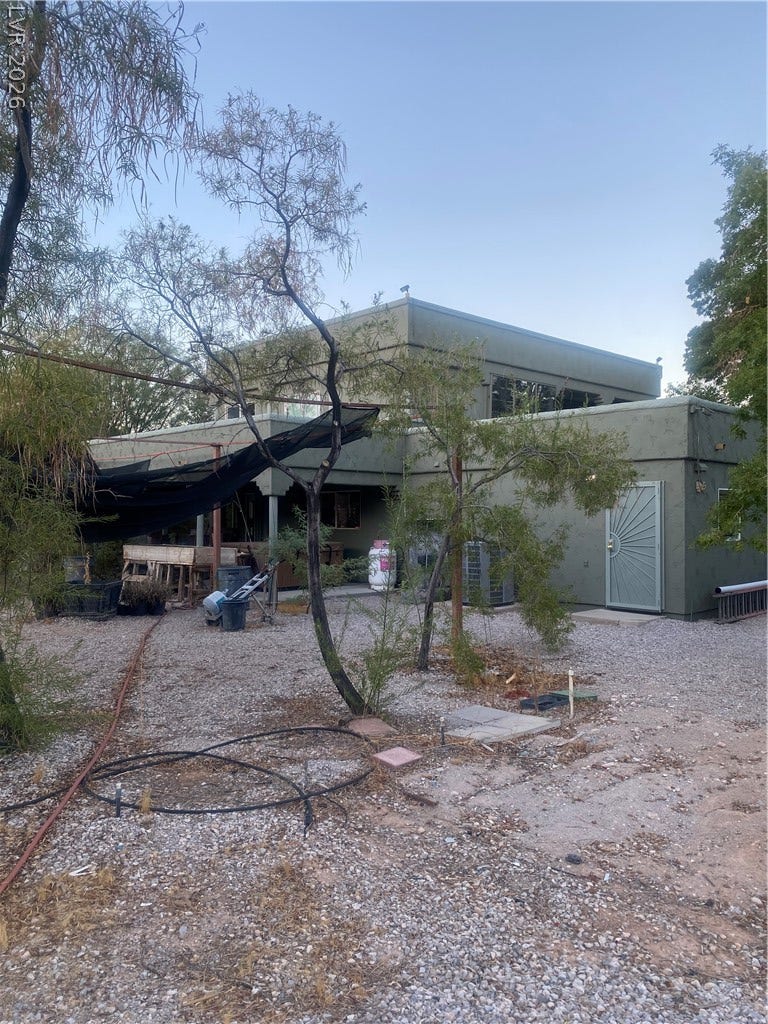 Mature trees shade the gravel yard behind a multi-level stucco home.