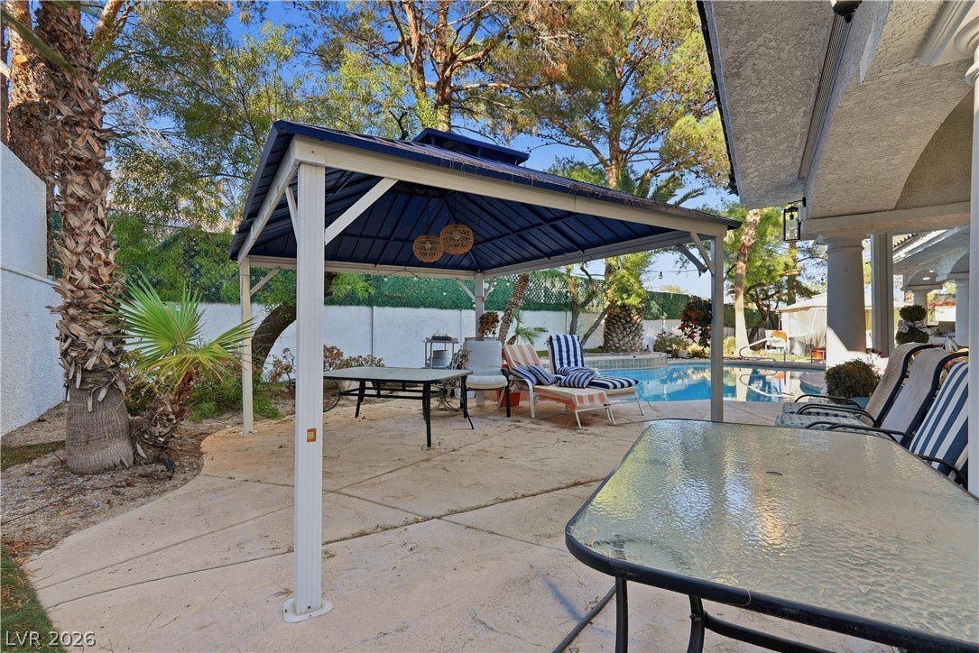 Mature trees frame a sun-drenched concrete patio featuring a shaded metal gazebo beside the pool.