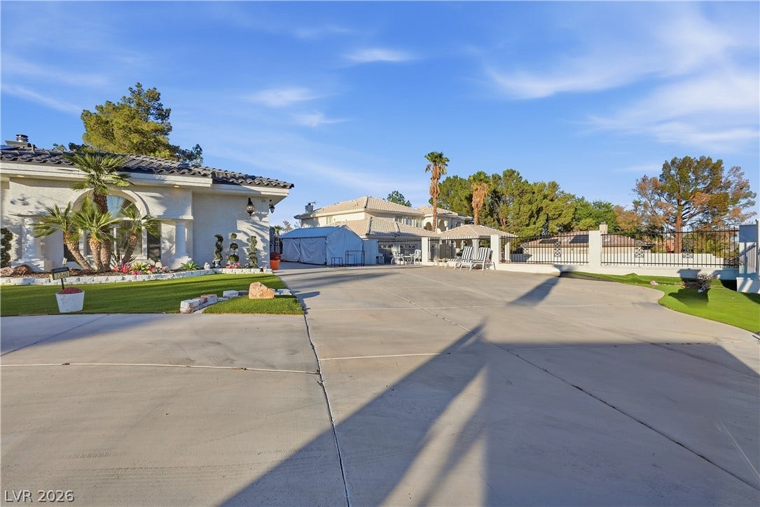 An expansive concrete driveway leads past landscaped grounds toward a sprawling residential estate.