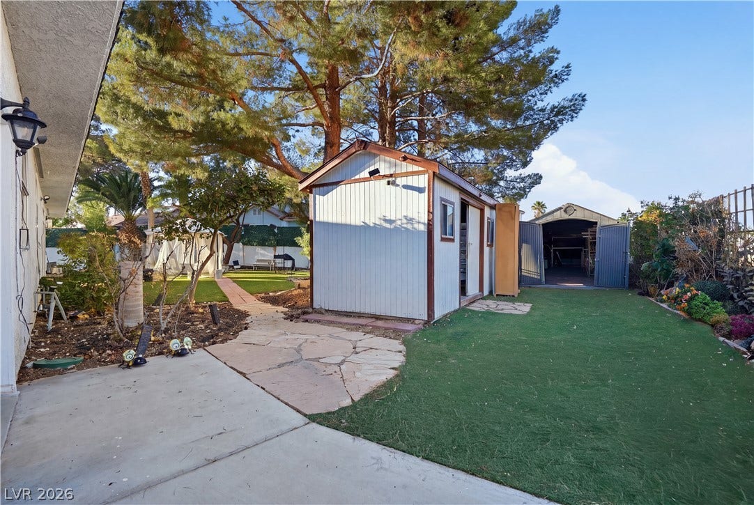 Mature trees shade a paved stone path leading past functional outbuildings in this spacious yard.