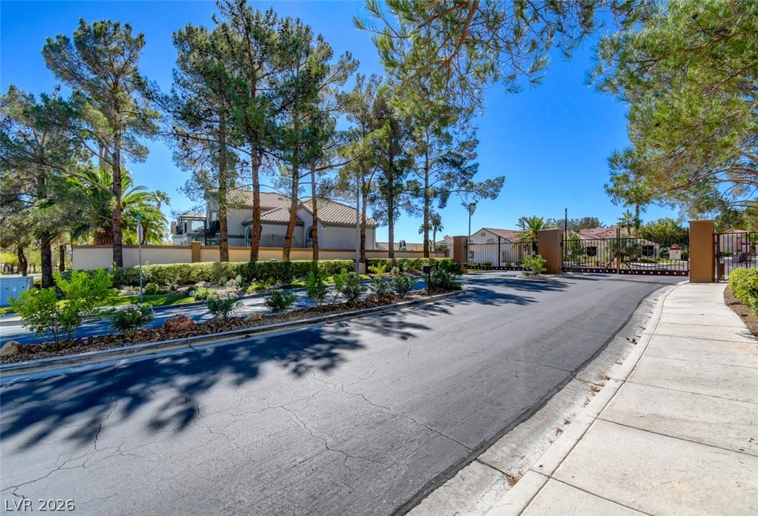 Mature trees line the paved entry street leading past secure gates under clear skies.