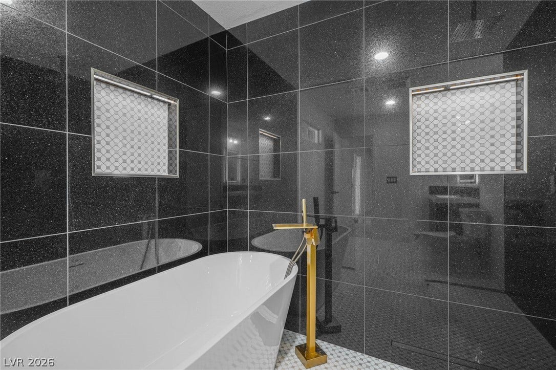 Dramatic dark wall tiles flank a modern soaking tub paired with a gold floor-mount filler.
