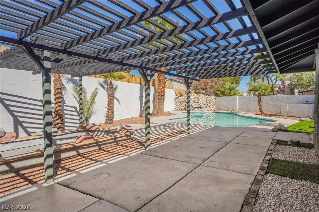 An inviting backyard oasis features a spacious concrete patio shaded by a large wooden pergola.
