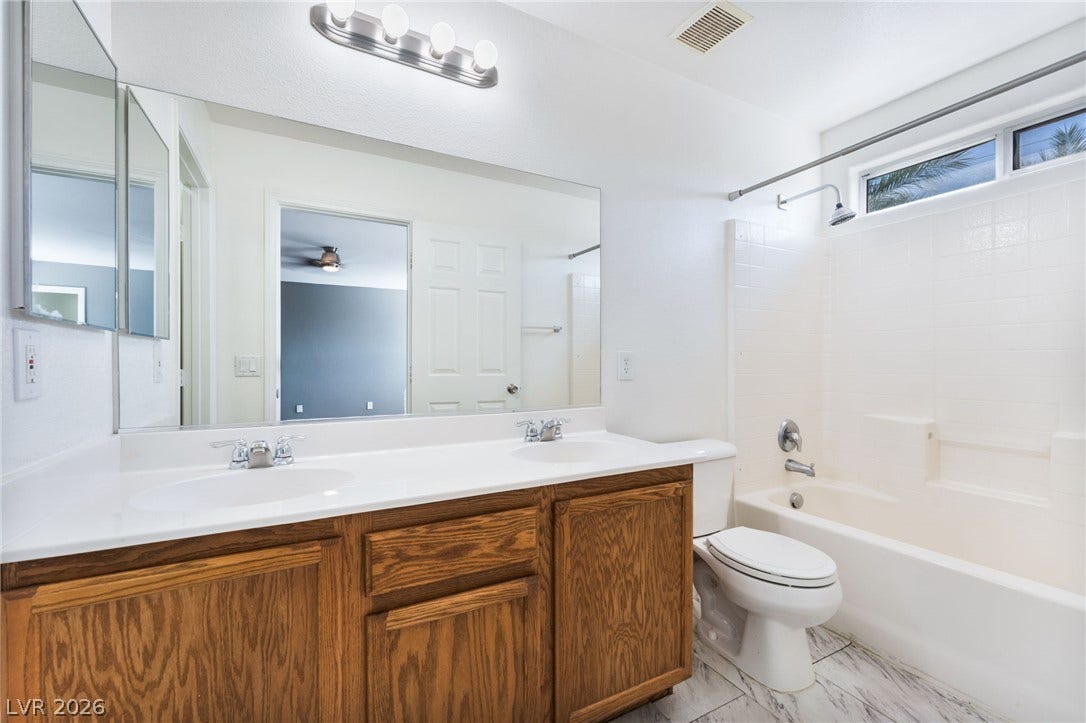 2825 Woodland Park Avenue, North Las Vegas, NV 89086 Natural light from a high window illuminates a classic bathroom featuring dual sinks and stone-patterned tile flooring.