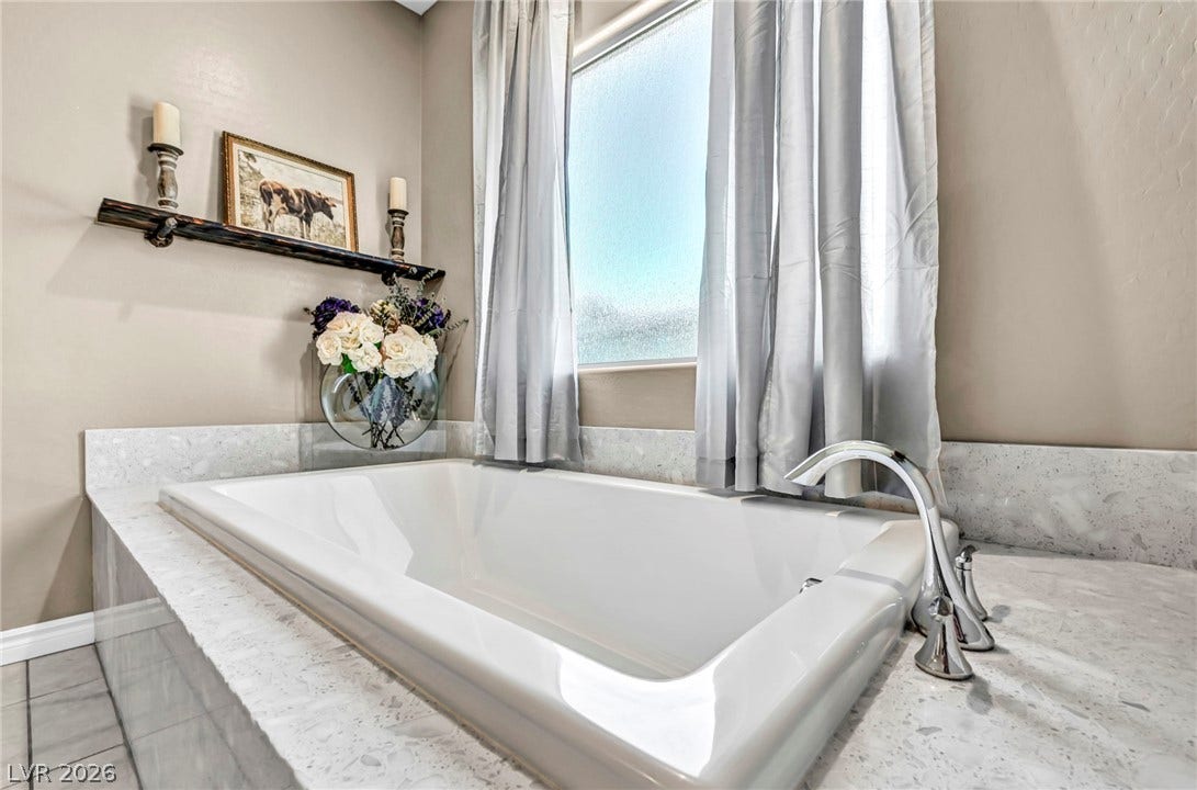 A deep soaking tub sits enveloped by classic stone surrounds beneath a bright picture window.