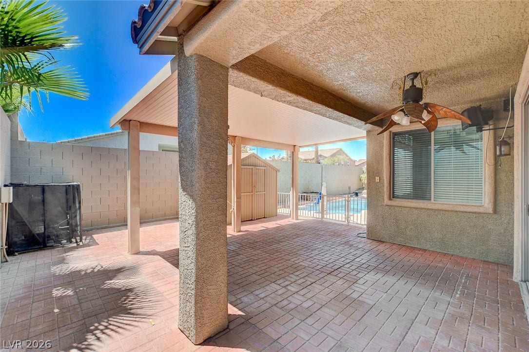 849 Ashford Way, Henderson, NV 89015 An inviting covered patio featuring brick pavers and a ceiling fan, overlooking a fenced pool area and shed.