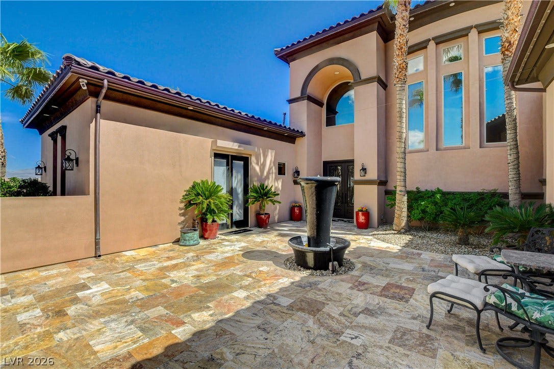 An inviting courtyard features warm stone pavers and a central water fountain under clear blue skies.