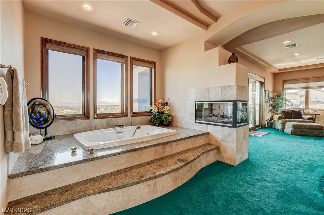 An elevated soaking tub features premium stone surrounds alongside a multi-sided fireplace and picture windows.