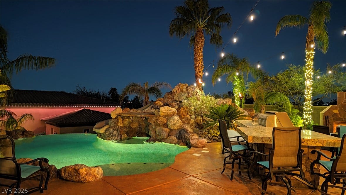 Lush palm trees and warm lighting frame a tropical swimming pool and outdoor kitchen.