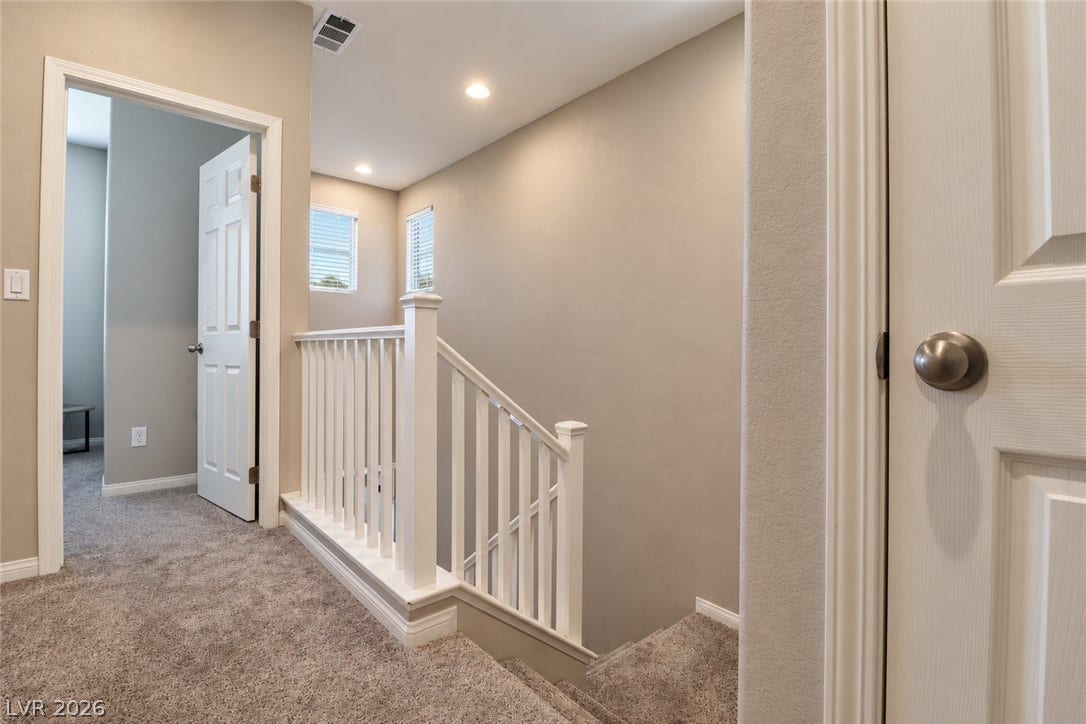 An upper-level landing showcases neutral tones, plush carpeting, and recessed lighting alongside the wooden staircase railing.