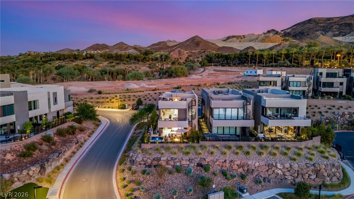 18 Heron Harbour Drive, Henderson, NV 89011 Modern multi-level homes nestle against a dramatic mountain backdrop under a sunset sky.
