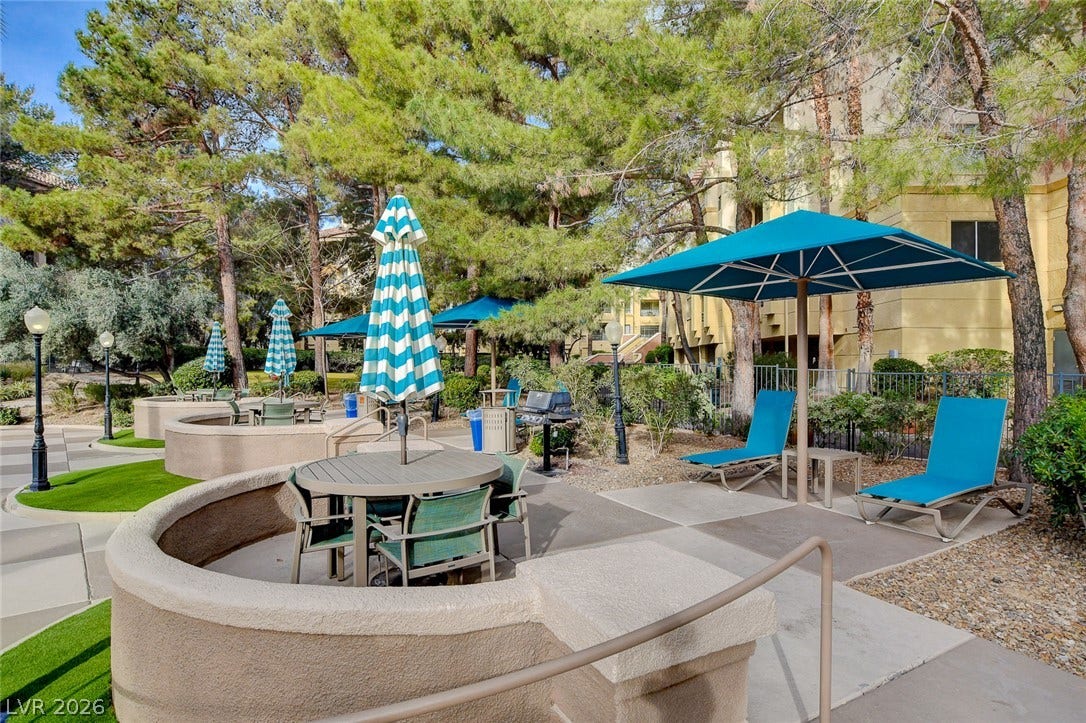 Mature trees surround a concrete patio seating area situated adjacent to residential buildings.