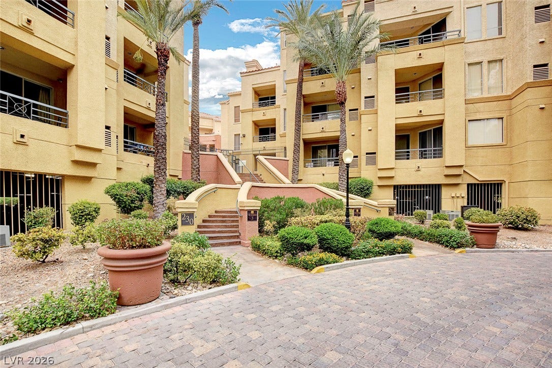 Multi-level stucco buildings flank a paved courtyard walkway framed by palm trees and manicured shrubbery.