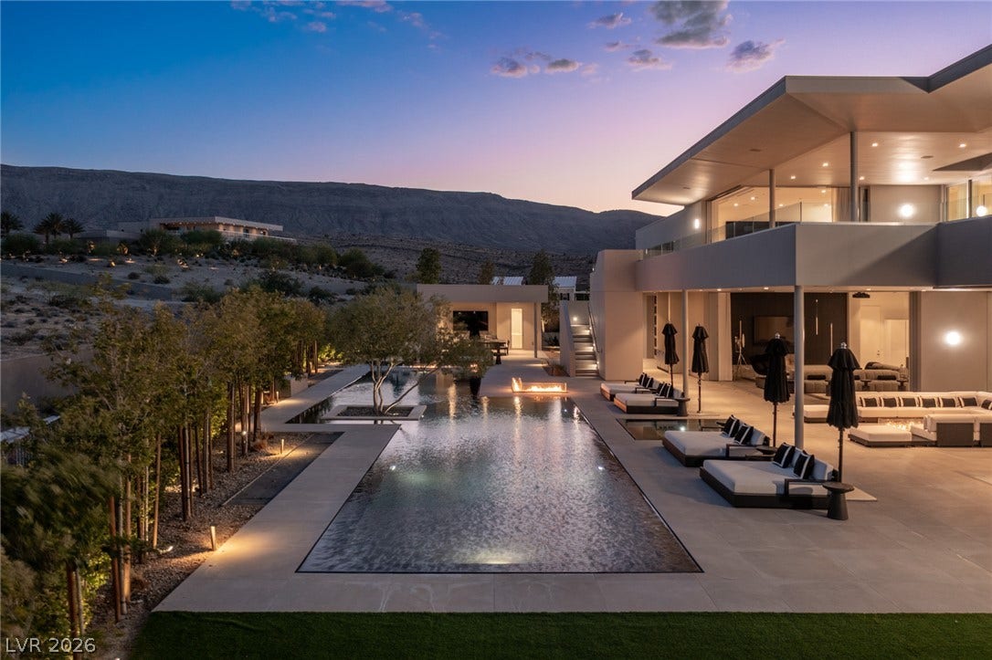 An illuminated desert oasis features a long swimming pool, fire pit, and ample patio seating under the twilight sky.
