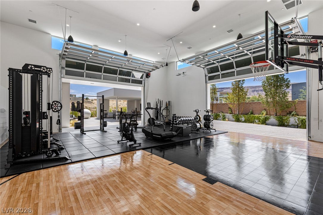 An indoor sports court with a basketball hoop and a full gym, featuring large glass garage doors that open to the outside.