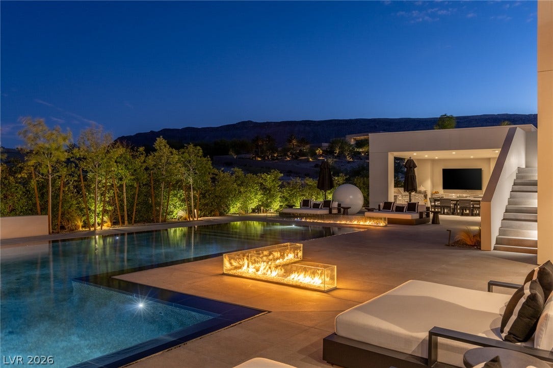 An illuminated backyard retreat boasts a shimmering pool and multiple fire features against a mountain view.