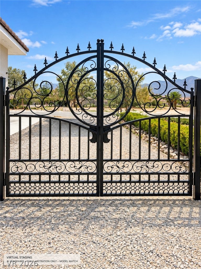 Virtually staged. 
An ornate wrought iron gate opens to a gravel driveway surrounded by natural desert landscaping.