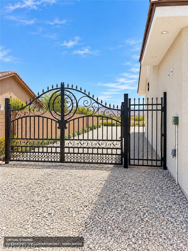 Virtually staged. 
An elegant wrought iron gate frames the entrance along a gravel driveway under clear skies.