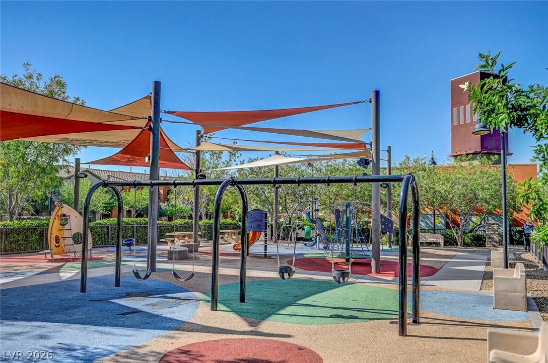 A vibrant playground features swings, climbing structures, and protective shade canopies on a sunny day