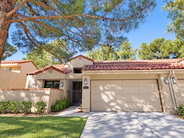 Mature trees shade the concrete driveway leading to a Spanish tile roof home.