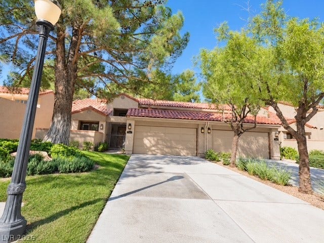 Mature trees shade the concrete driveway leading to a multi-car garage with Spanish tile roofing.