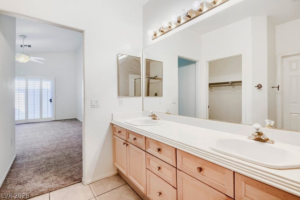 6729 Quarterhorse Lane, Las Vegas, NV 89148 Spacious primary bathroom featuring a dual-sink vanity with generous wood cabinetry and drawer storage, expansive wall mirror, vanity lighting, neutral tile flooring, and a walk-in closet conveniently located just off the bath.