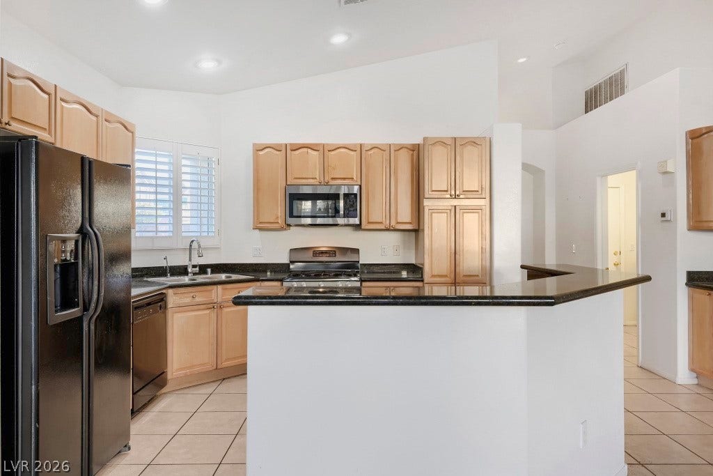 6729 Quarterhorse Lane, Las Vegas, NV 89148 Spacious kitchen featuring a large wraparound granite breakfast bar, abundant wood cabinetry, double-basin sink, recessed lighting, plantation shutters, and appliances including side-by-side refrigerator, dishwasher, stainless steel gas range, and built-in microwave.