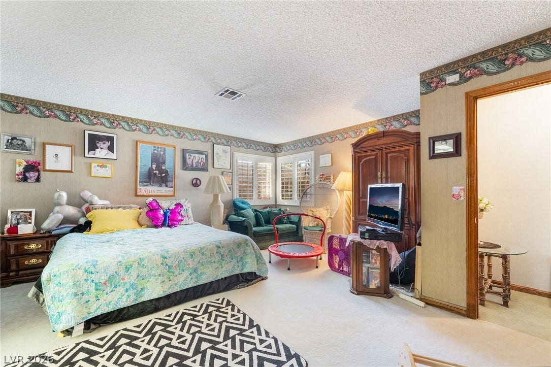 A spacious bedroom features neutral carpeting, decorative wall borders, and plantation-style window shutters.