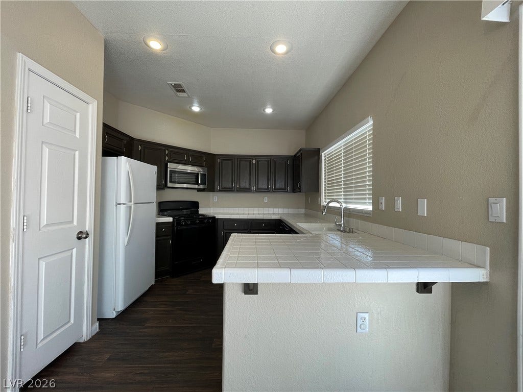 7873 Blue Brook Drive, Las Vegas, NV 89147 Dark cabinetry and classic tile countertops define this functional kitchen workspace with wood-look flooring.